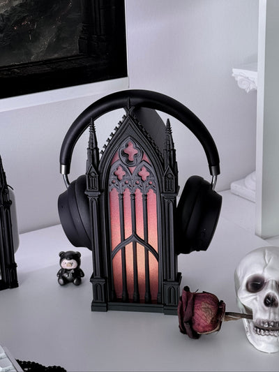 Black gothic-style goth headband headphones displayed on a decorative black gothic arch lamp with a glowing pink and orange gothic window design, set on a white desk with skull and gothic decor props.