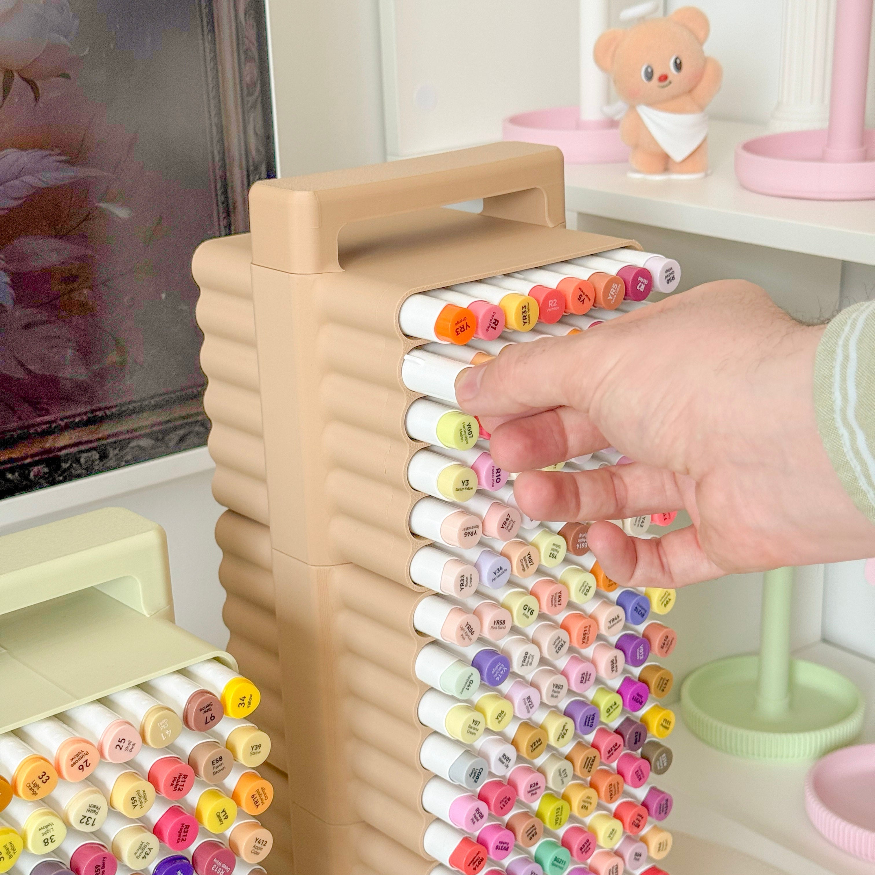 Colorful markers on a holder with a hand selecting a marker, set against a blurred indoor background.